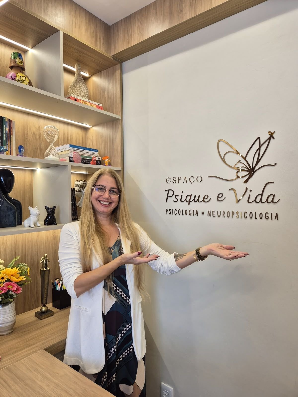 Viviane Gatis em pé no consultório, apontando para o letreiro do Espaço Psique e Vida na parede.
